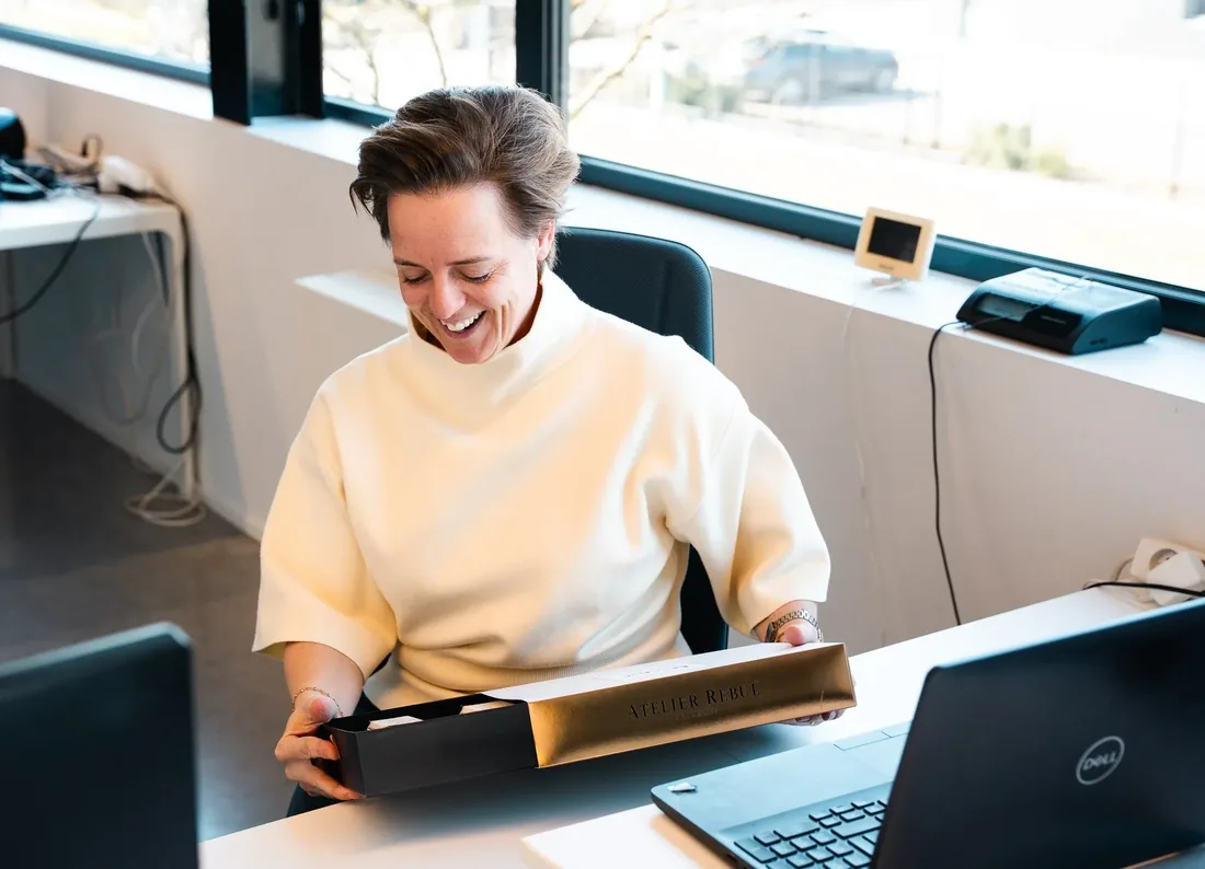 Een medewerker opent aan haar bureau een geschenkbox met een gouden deksel.